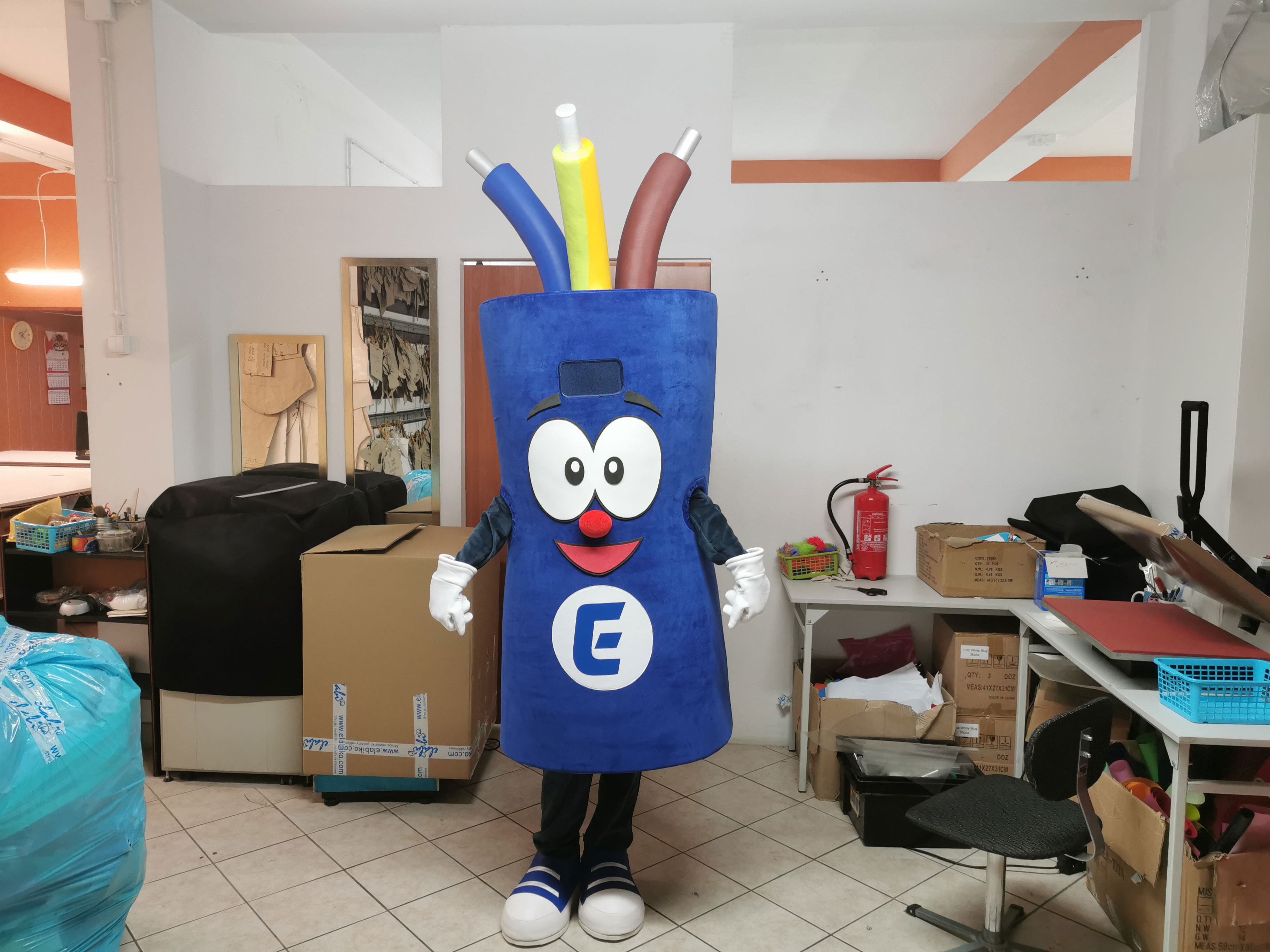 Na fotografii widnieje kostium reklamowy kabel, pozujący do zdjęcia w jednym z pomieszczeń produkcyjnych firmy Elabika. Stąd wokół widać typowe sprzęty dla projektowej pracowni krawieckiej